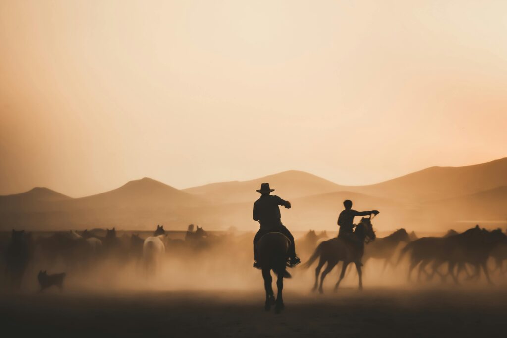 Silhouetted cowboys herding horses across a dusty landscape during a stunning sunset.